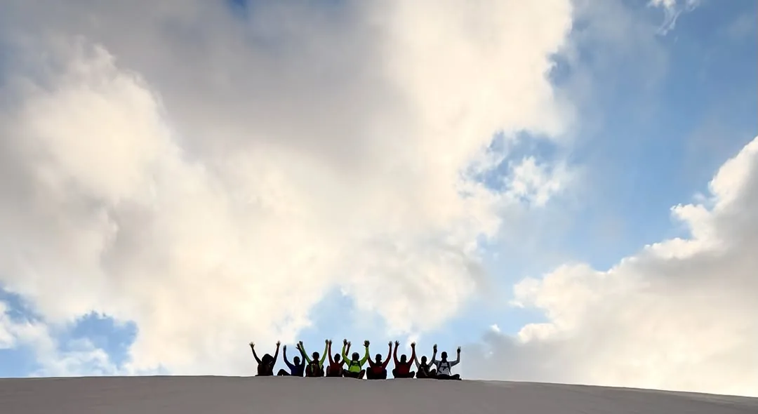 O grupo da travessia sentado no alto de uma duna branca, de braços erguidos contra o céu com nuvens