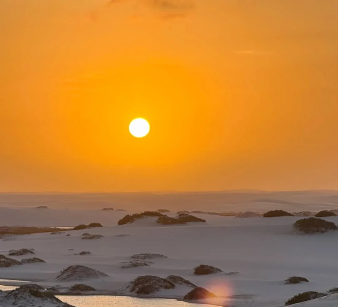 Sol baixo e alaranjado sobre as dunas e uma lagoa dos Lençóis Maranhenses