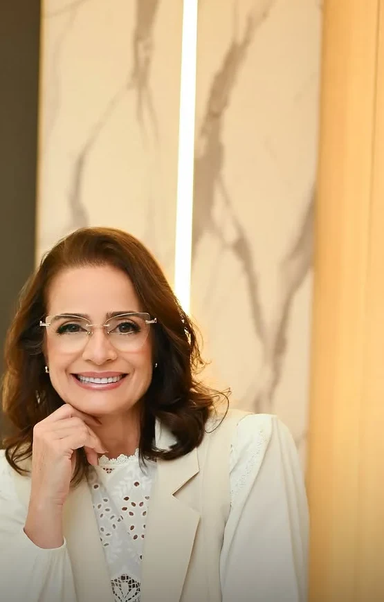 Dra. Luciana Maldonado, de blazer branco, sorrindo com a mão no queixo diante do mármore da recepção