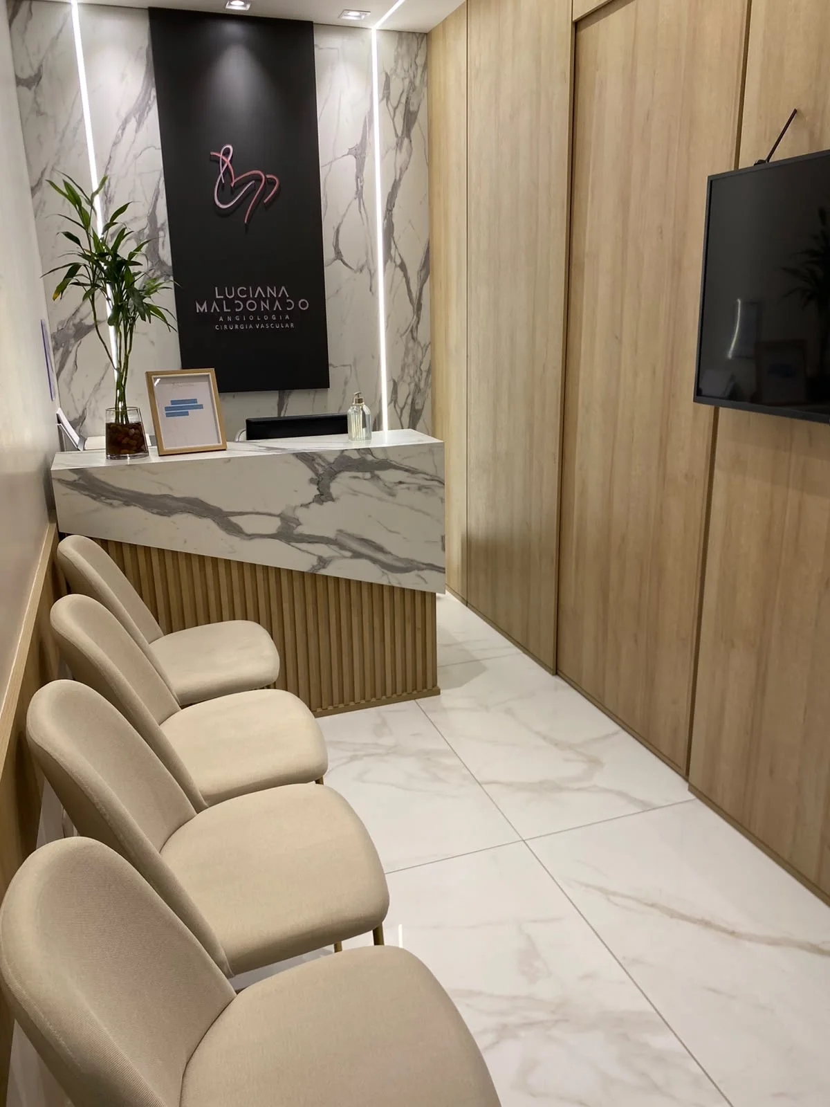 Sala de espera com poltronas claras enfileiradas, balcão de mármore e, ao fundo, o painel grafite com o logo