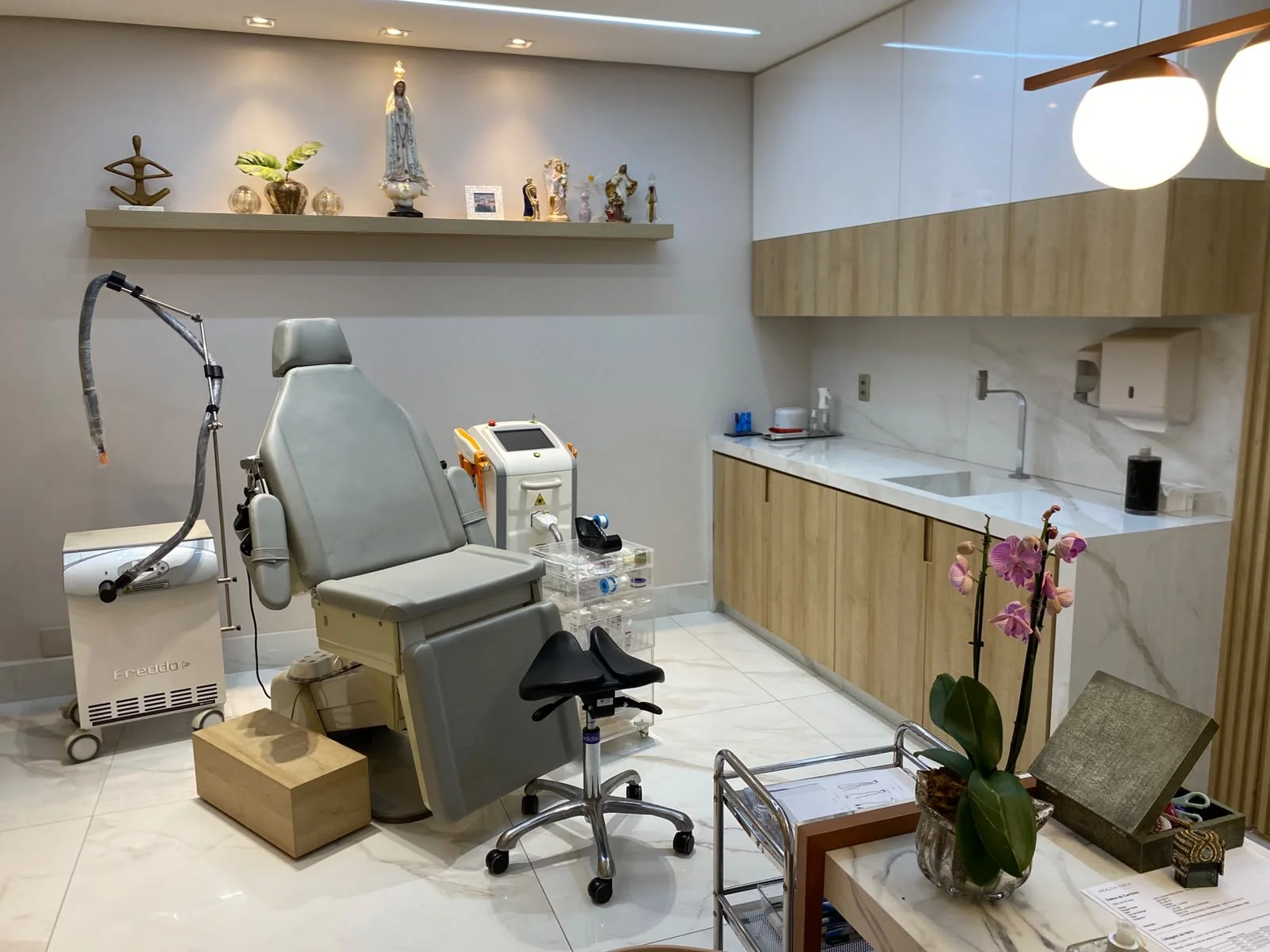 Sala de procedimentos com cadeira clínica, aparelho de laser, bancada de madeira clara e imagens de santos na prateleira