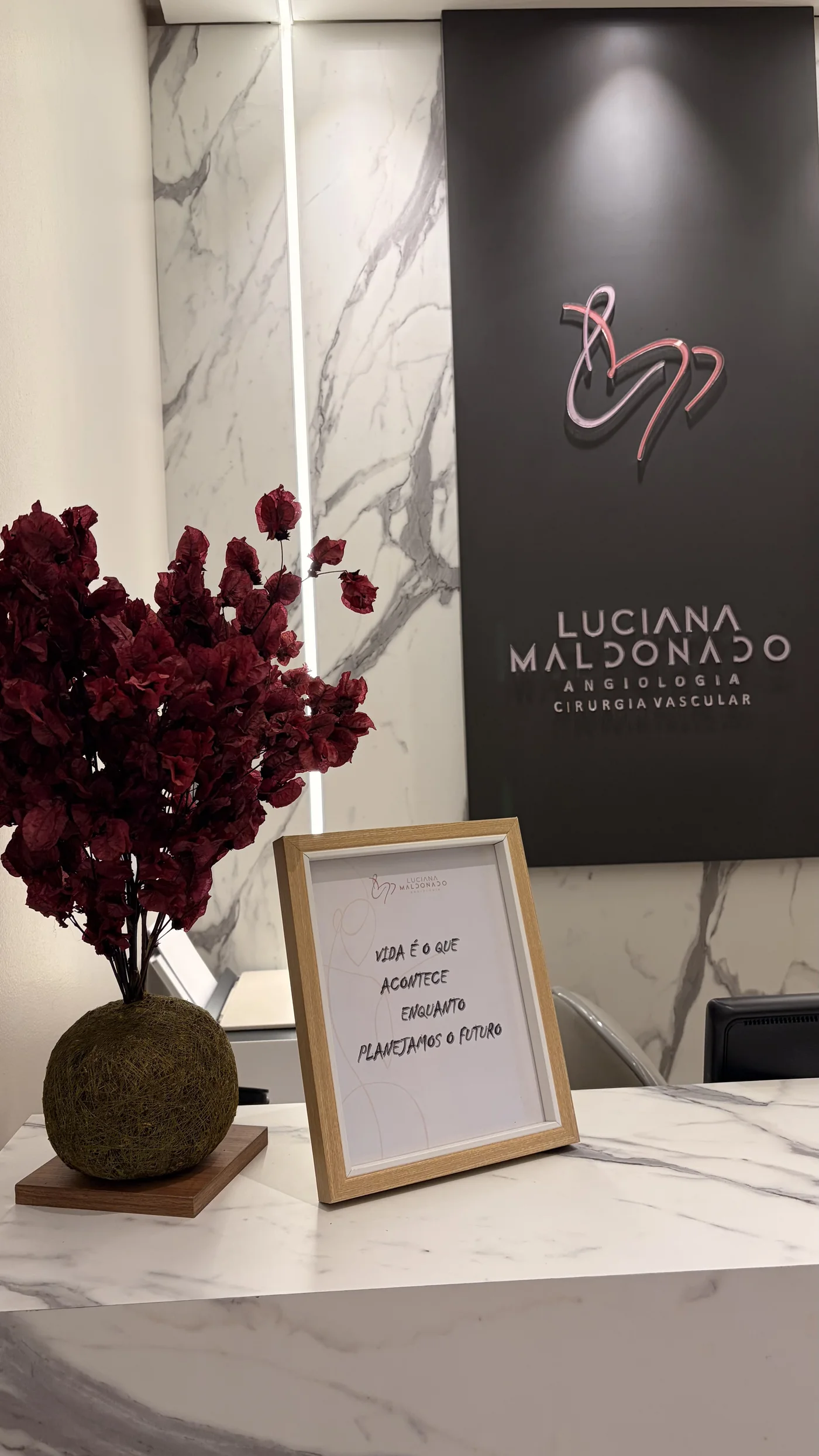 Recepção do consultório: painel grafite com o logo rosé iluminado, parede de mármore branco com veios cinza, flores bordô e um quadro sobre o balcão de mármore