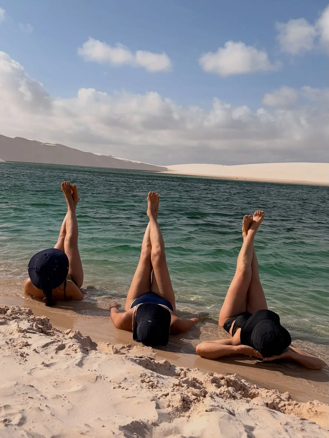 Três mulheres deitadas na areia à beira de uma lagoa dos Lençóis Maranhenses, com as pernas para o ar e chapéus cobrindo o rosto