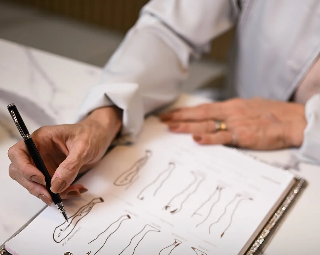 A mão da Dra. Luciana, de caneta, desenhando o contorno de pernas num caderno sobre a mesa de mármore
