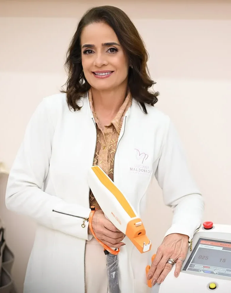 A Dra. Luciana, de jaleco branco, sorrindo, com a peça do laser na mão ao lado do aparelho